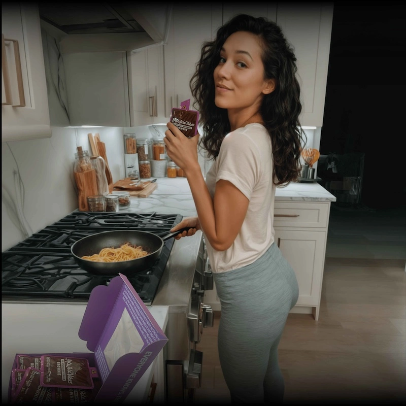 Woman with curly hair cooking pasta in modern white kitchen while holding BakeSale Vibes product, with additional packages visible on marble countertop