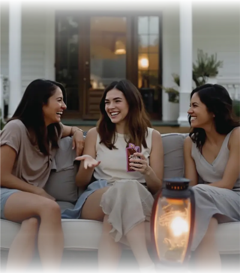 Three women friends laughing and socializing while enjoying BakeSale Vibes product on elegant outdoor patio with warm lighting
