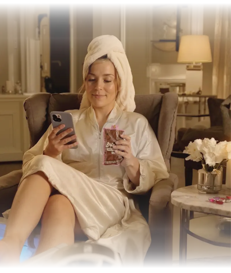 Woman in white bathrobe and towel relaxing in luxurious chair while enjoying BakeSale Vibes product and using phone in elegant spa-like setting